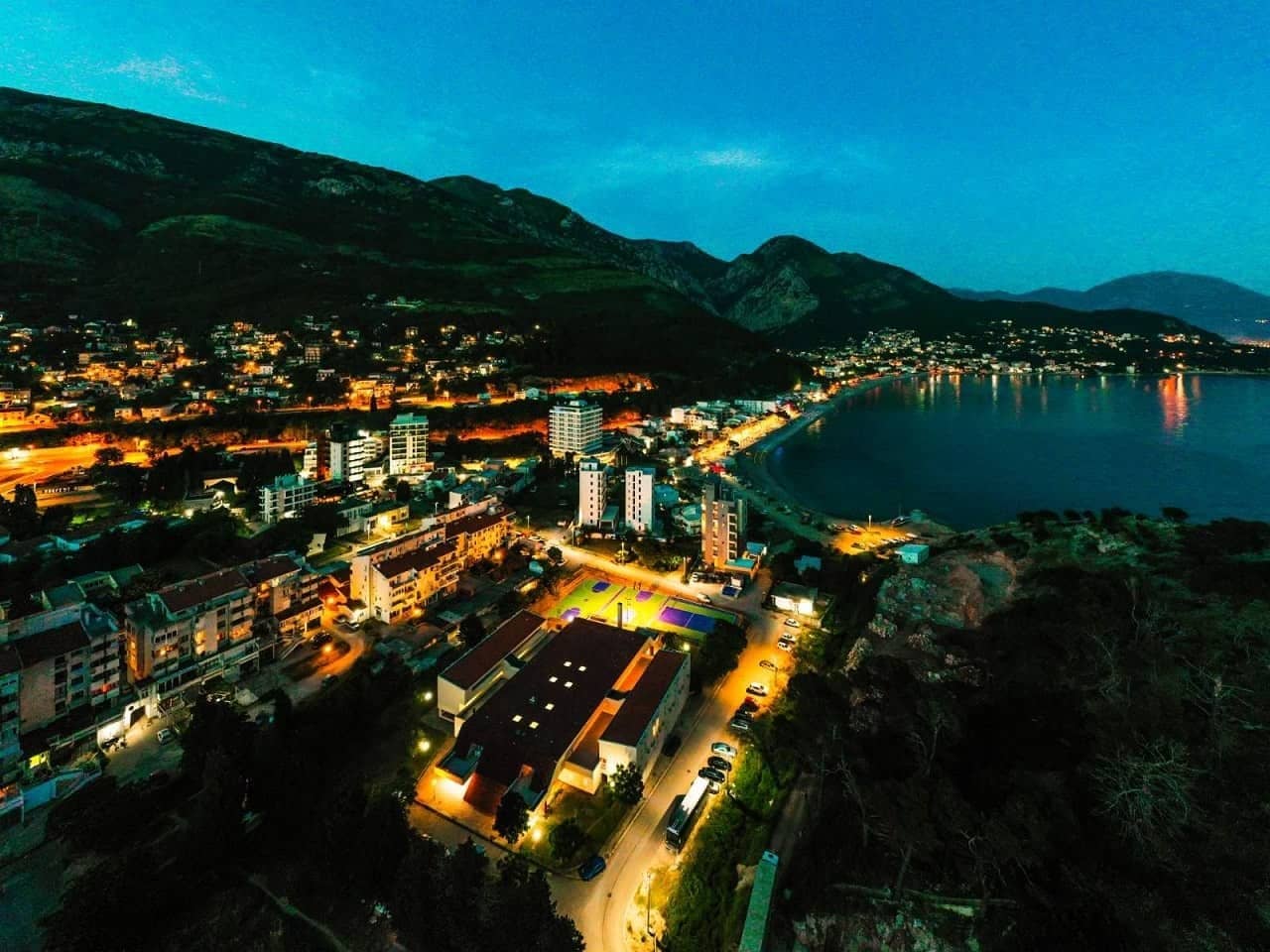 Sutomore u sumrak iz vazduha — zaliv, svjetla grada i osvijetljeni košarkaški tereni uz smještaj